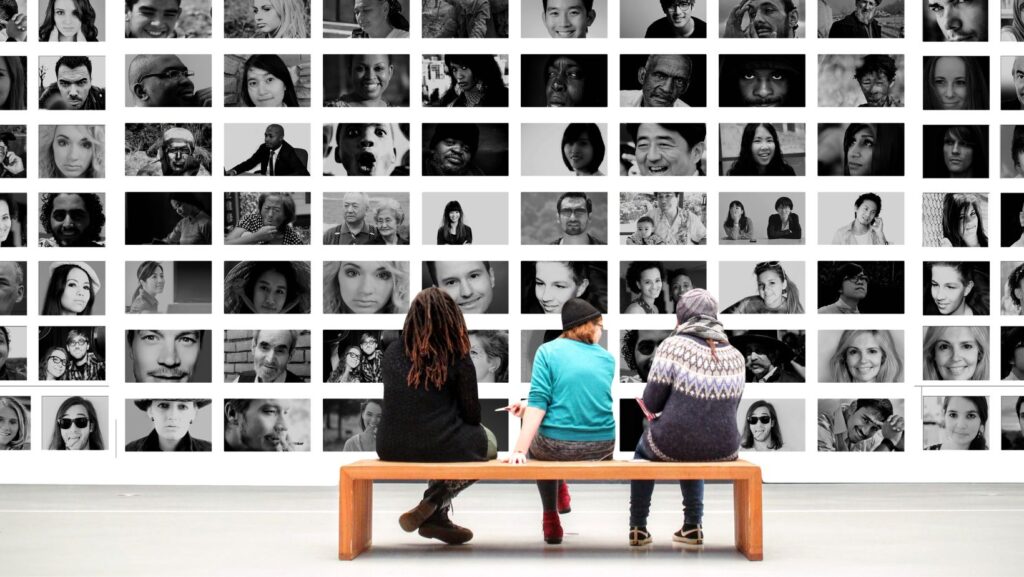 Three people are sitting on a bench facing away from the viewer, observing a wall collage of pictures. The pictures feature individuals of various ages and backgrounds, showcasing diversity. The images are in black and white, except for the bench and the three individuals sitting on it. The scene resembles people observing artwork at a museum.