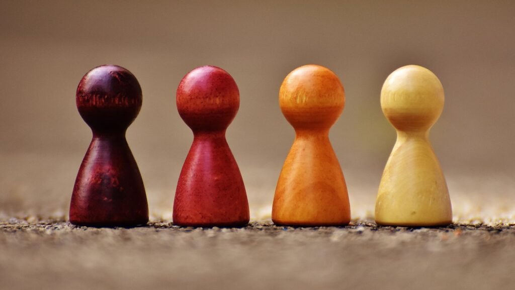 Four modern-looking chess pieces of the same shape and size stand side by side on the ground. They are colored maroon, red, orange, and yellow, symbolizing diversity. The background is beige. The image represents diversity through colorful chess pieces in a minimalist setting.